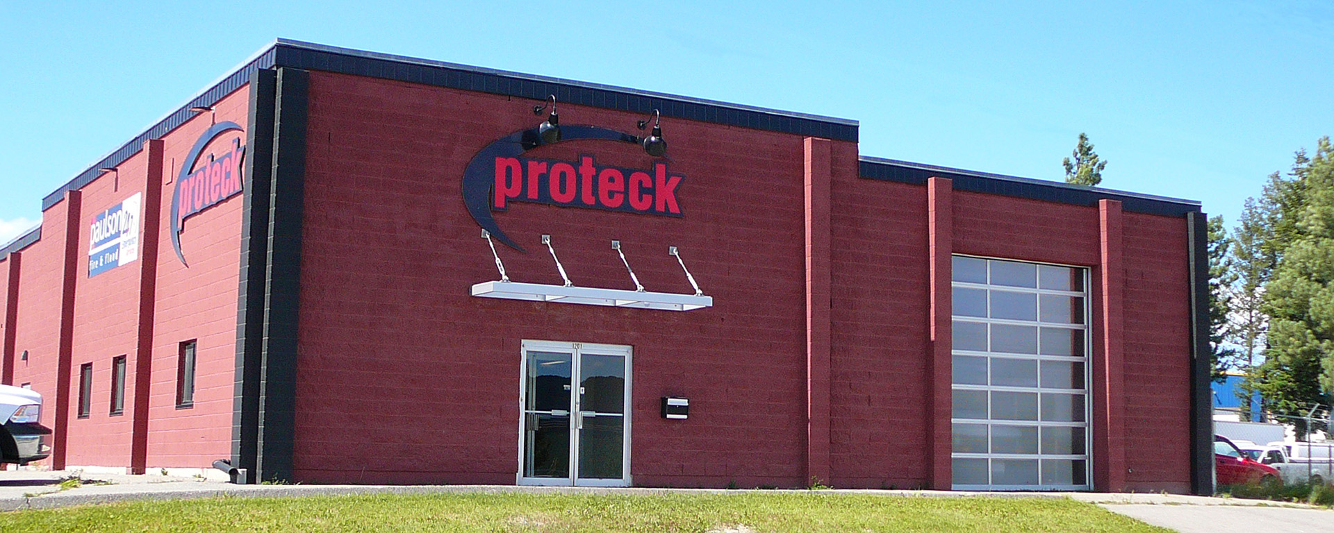 Outside of Proteck building. Building is red brick and has a large glass-window overhead door. 