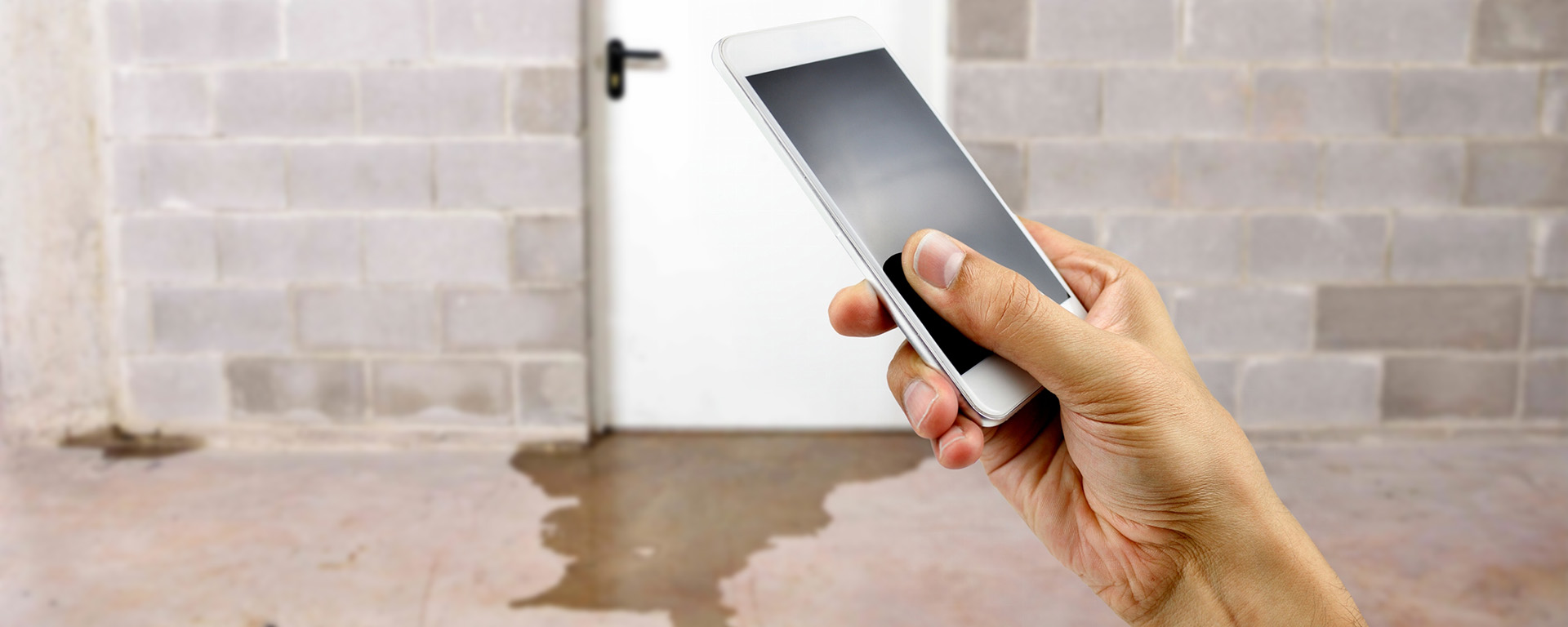 A hand holding up a smart phone with water damage in basement caused by sewer backflow due to clogged sanitary drain. 