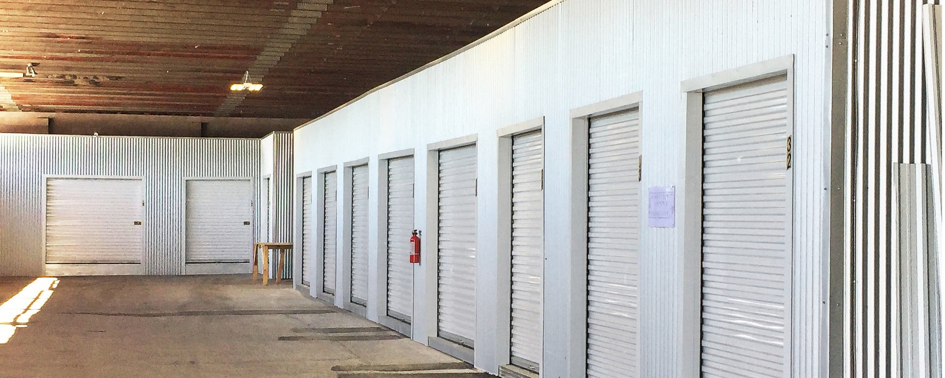 Interior of First Avenue Self Storage featuring a clean, well-lit hallway with a row of white roll-up storage units and a fire extinguisher. 