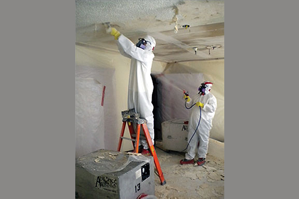 Man on a ladder scraping the ceiling while another man uses a sprayer